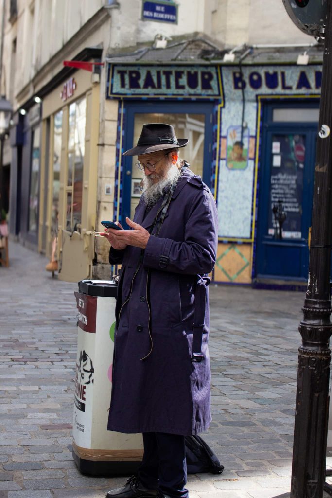 homme habillé dun lon trench bleu marine, et d'un chapeau noir, regardant son telephone dans les rues de paris, portant une longue barbe blanche