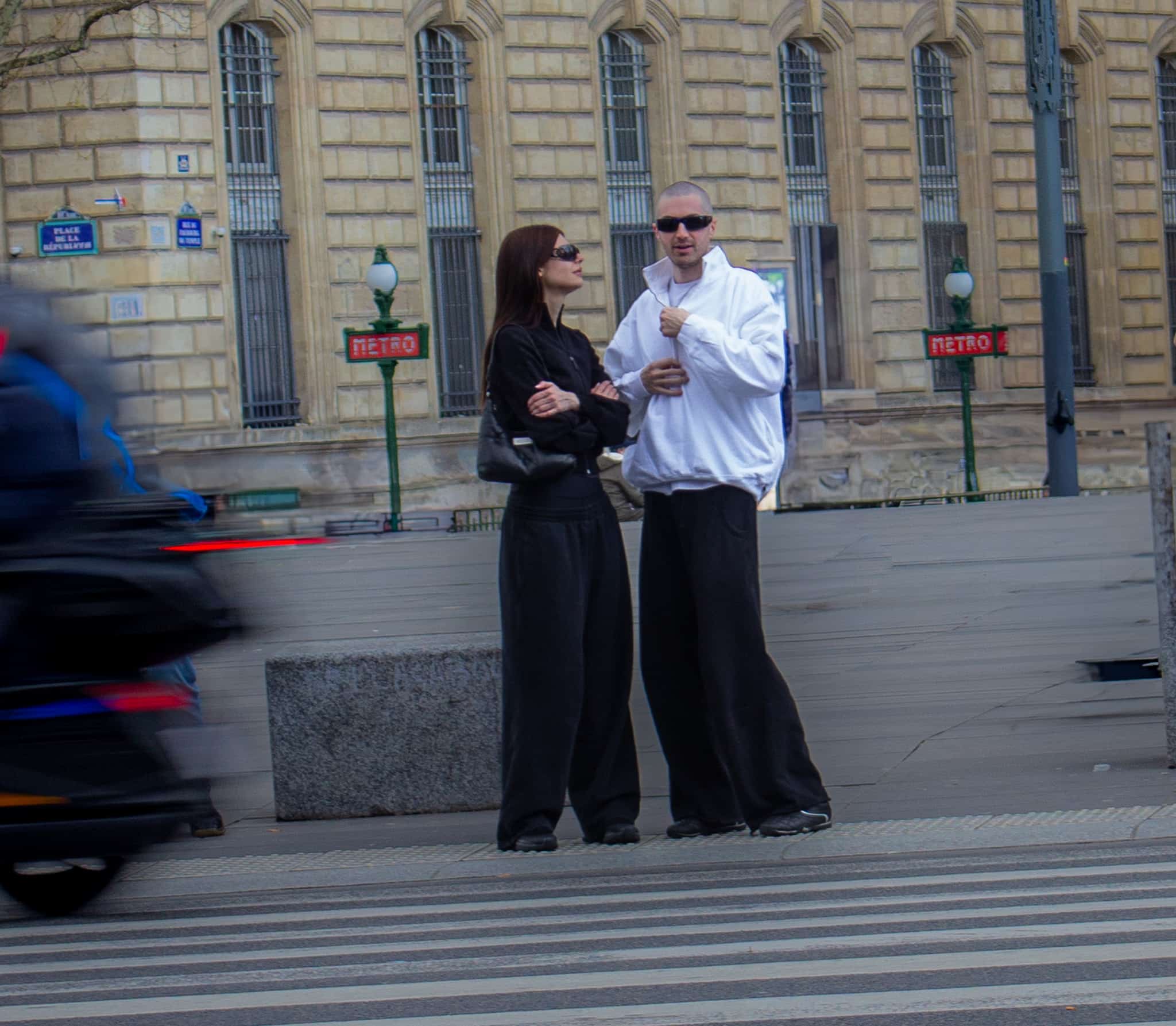 une jeune couple attendant de traverser à un passage piéton. l'homme porte une vest blanc et un pantalon noir, et la femme est tout de noir vêtue. Les deux sont dans un style tech wear, type techno